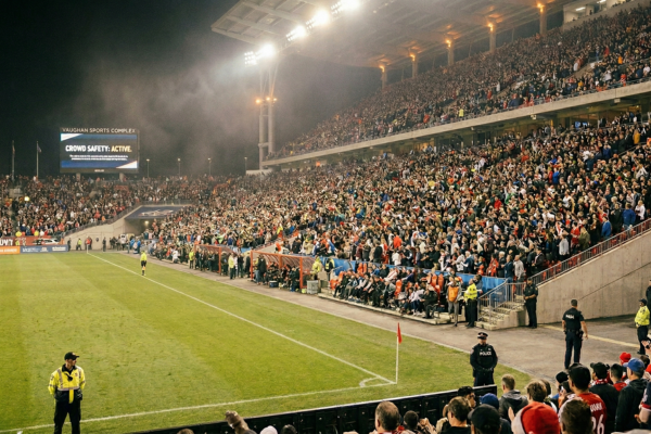 Professional event security personnel monitoring a massive crowd of fans from the field level of a packed, brightly lit soccer stadium at night